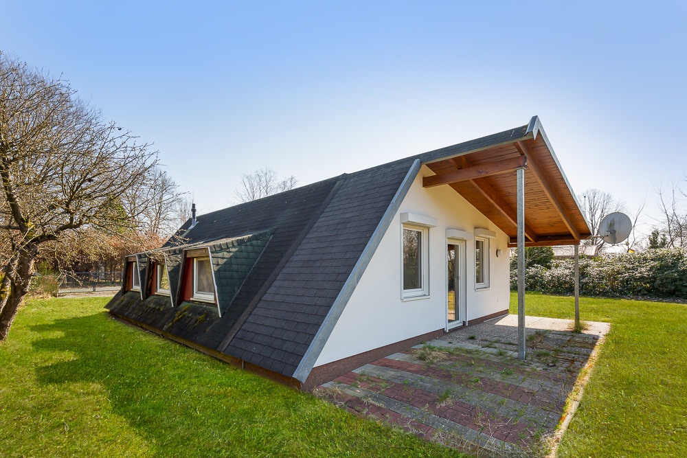 Haus mit schrägem Dach, weißen Wänden, Terrasse und großem Garten bei klarem Himmel.