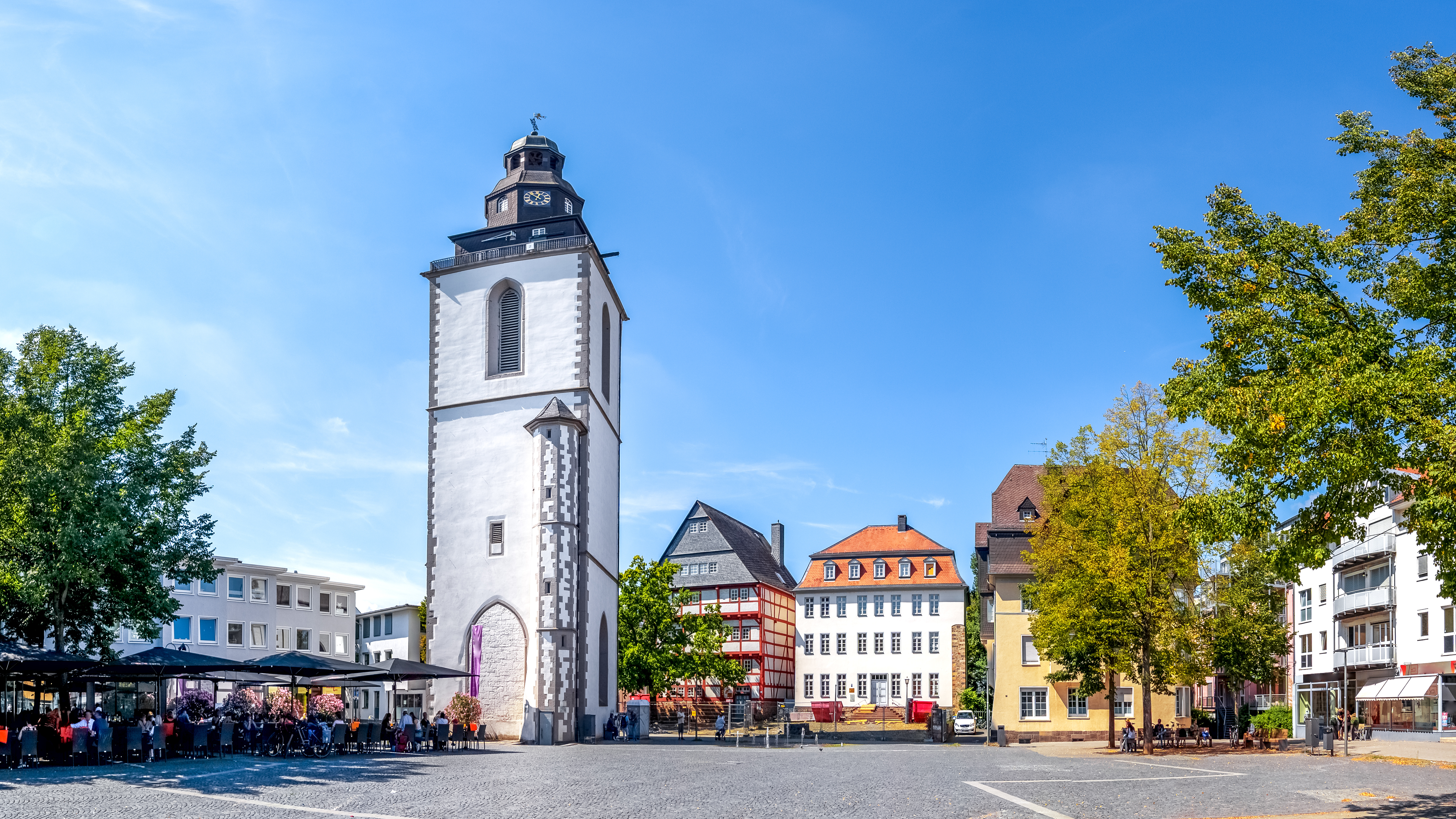 Ein Marktplatz mit einem weißen Turm, Bäumen und mehreren Wohngebäuden im Hintergrund.