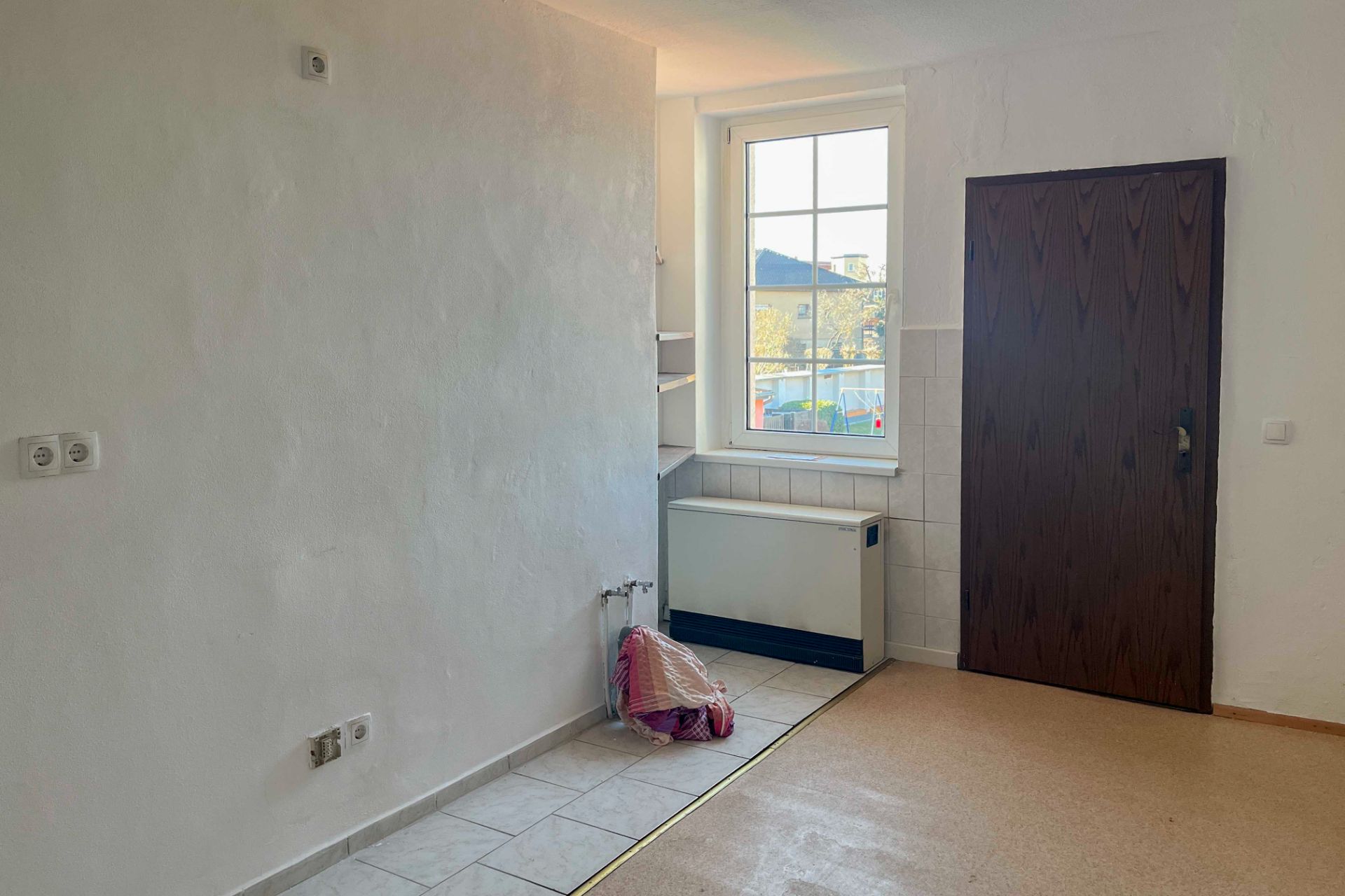 Heller Raum mit weißer Wand, Fenster, brauner Tür und Heizung unter dem Fenster.