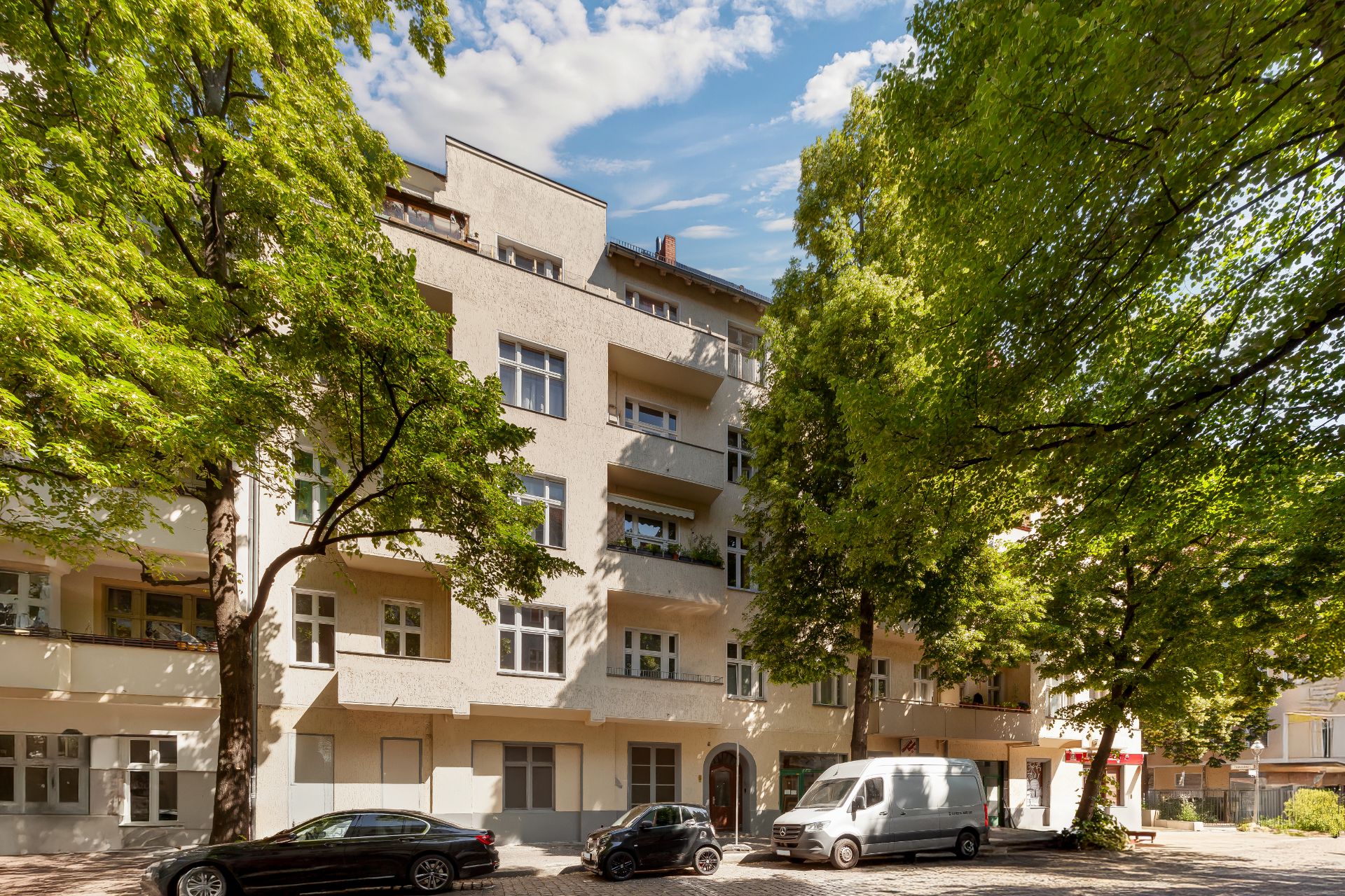 Mehrstöckiges Wohnhaus mit Balkonen und großen Bäumen davor, geparkte Autos an der Straße.