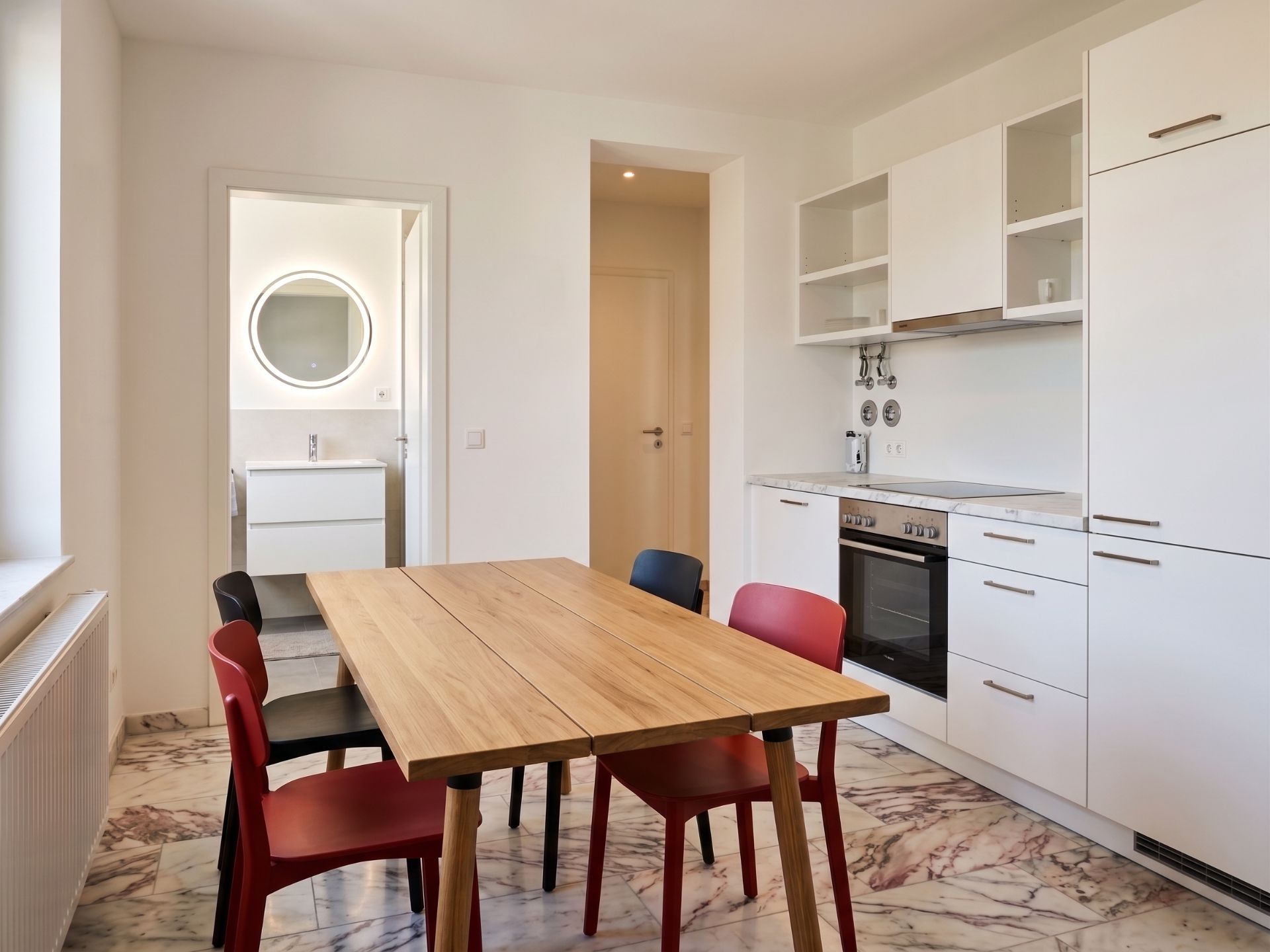 Moderne Küche mit Holztisch, bunten Stühlen und Zugang zu einem Bad mit rundem Spiegel.