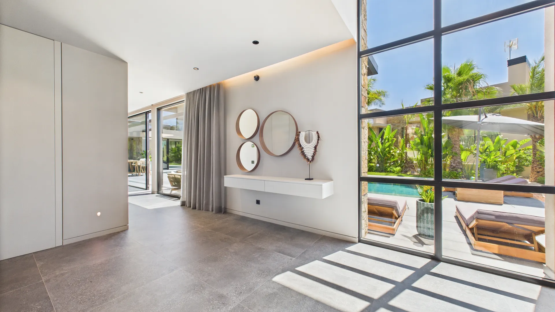 Helles Wohnzimmer mit großen Fenstern, Spiegeln an der Wand und Blick auf einen Garten mit Pool.
