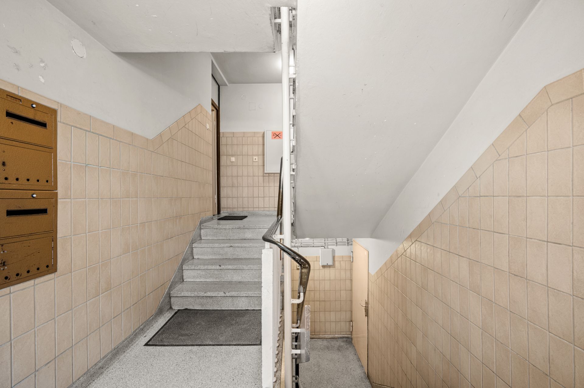 Treppenhaus mit beigen Fliesen, Briefkästen und grauen Stufen in einem Wohnhaus.