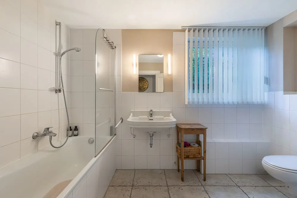 Helles Badezimmer mit Badewanne, Waschbecken, Spiegel und Fenster mit Lamellenvorhang.