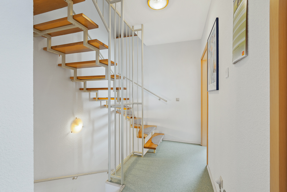 Heller Flur mit offener Holztreppe und weißen Wänden in einem Wohnhaus.