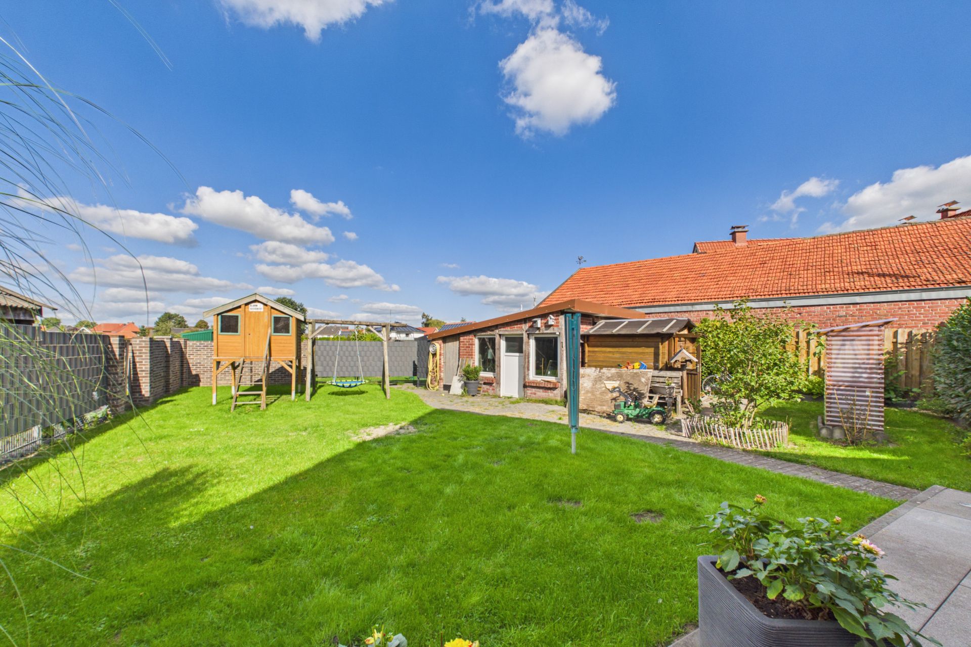 Grüner Garten mit Rasen, Spielhaus, Schaukel und Haus mit rotem Dach im Hintergrund.