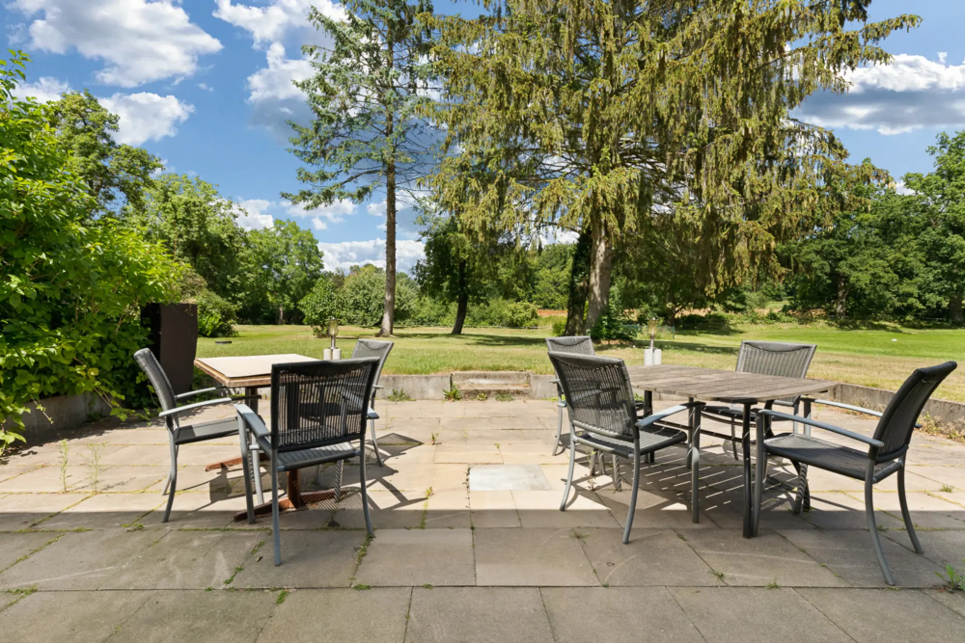 Garten mit Terrasse, mehreren Stühlen und Tischen, umgeben von Bäumen und Rasen.