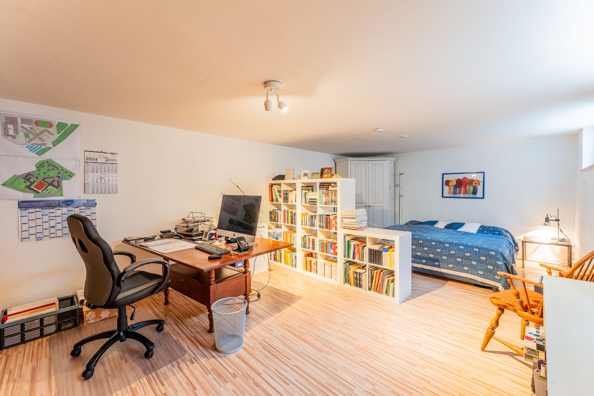 Helles Zimmer mit Schreibtisch, Bücherregal und Bett, aufgeteilt in Arbeits- und Schlafbereich.