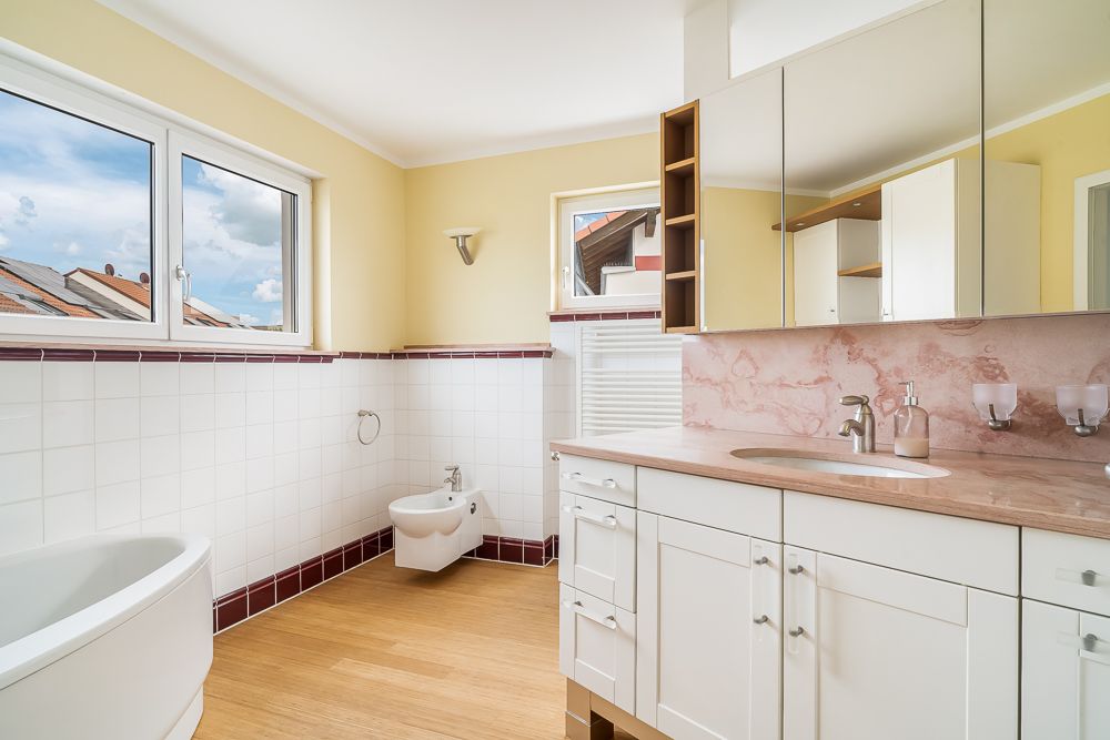 Helles Badezimmer mit Badewanne, Bidet, großem Spiegel und Waschbecken, Holzboden und Fenstern.