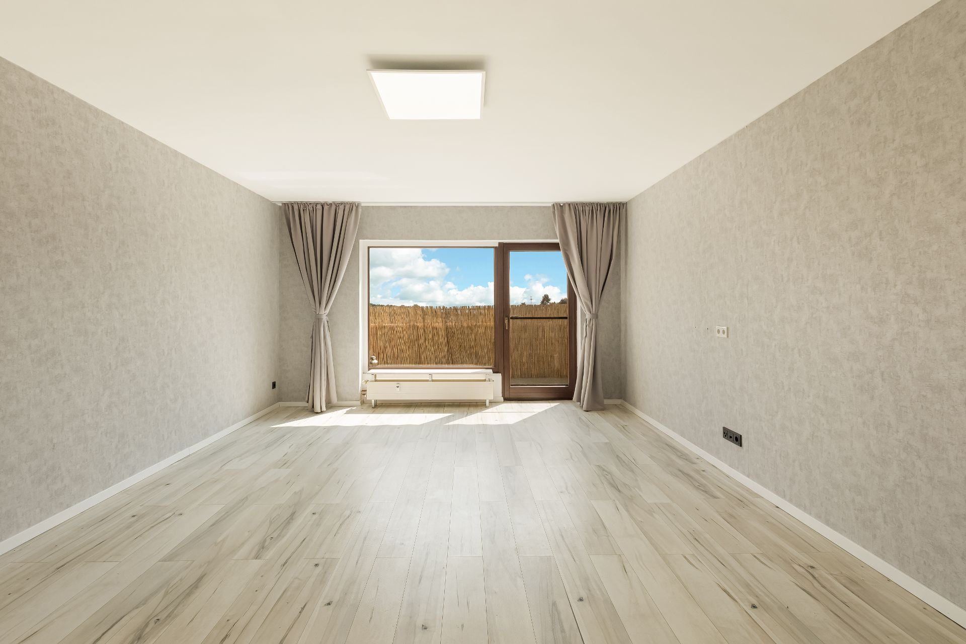 Helles, leeres Wohnzimmer mit großen Fenstern, Vorhängen und hellem Holzboden.