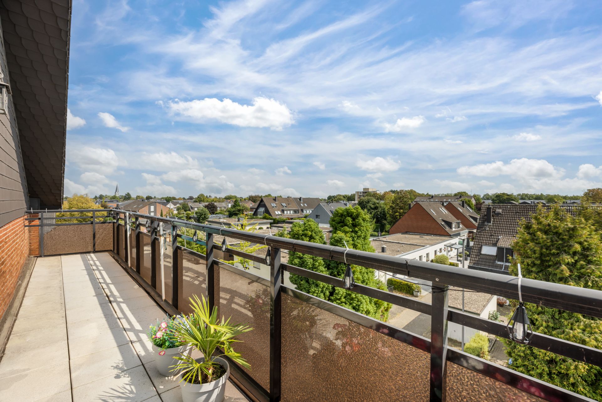 Großer Balkon mit Pflanzen und Ausblick auf Wohnhäuser und Bäume unter blauem Himmel.