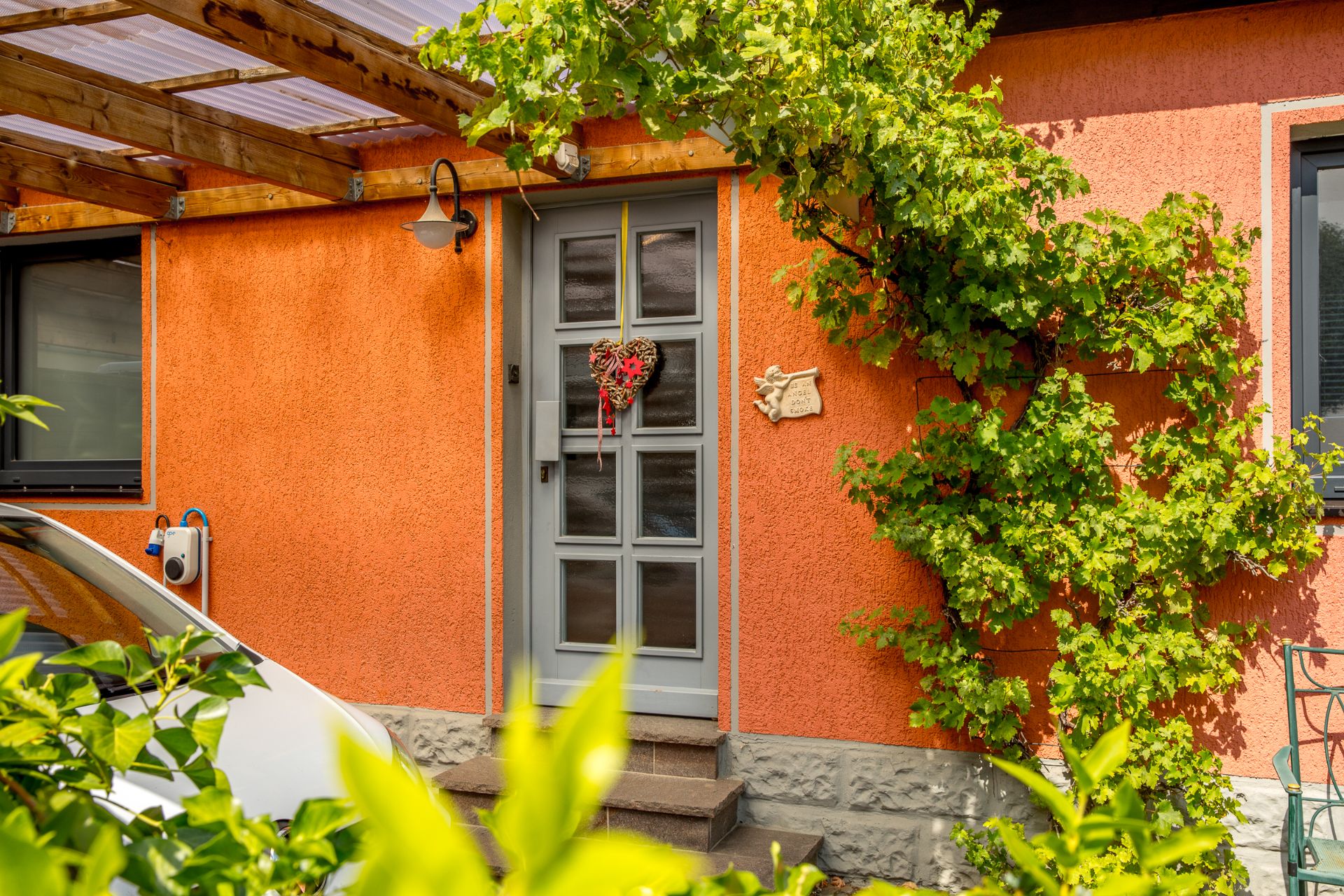 Eingangstür eines Hauses mit orangefarbener Wand, Herzdeko und grüner Kletterpflanze.