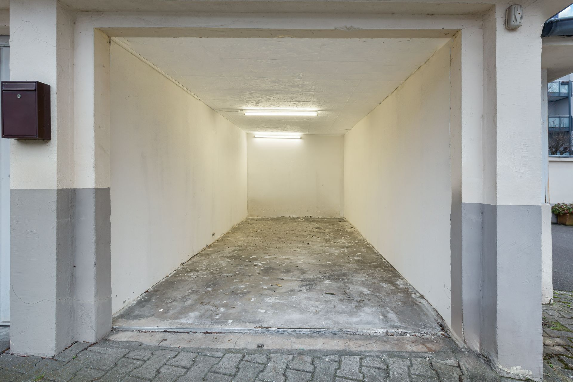 Leere Garage mit weißen Wänden und Betonboden, beleuchtet durch zwei Deckenlampen.