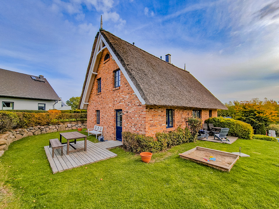 Backsteinhaus mit Reetdach, Holzterrasse, Sitzbank, Tisch, Sandkasten und Garten.