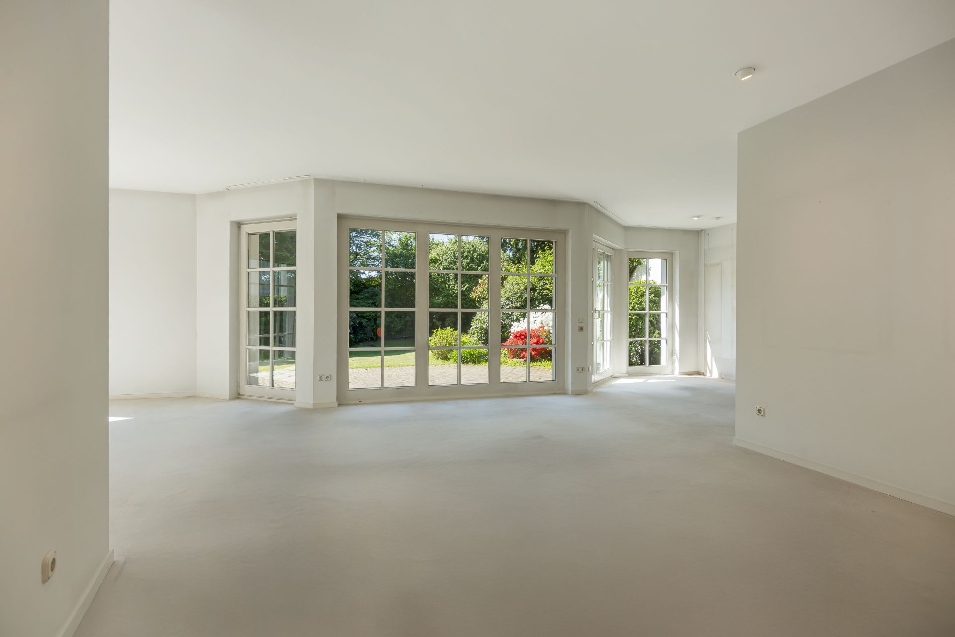 Heller, leerer Raum mit großen bodentiefen Fenstern und Blick in einen Garten.