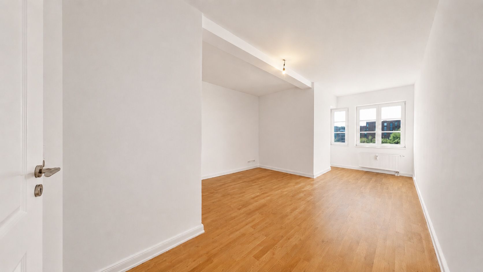 Heller, leerer Raum mit Holzfußboden, weißen Wänden und drei Fenstern.