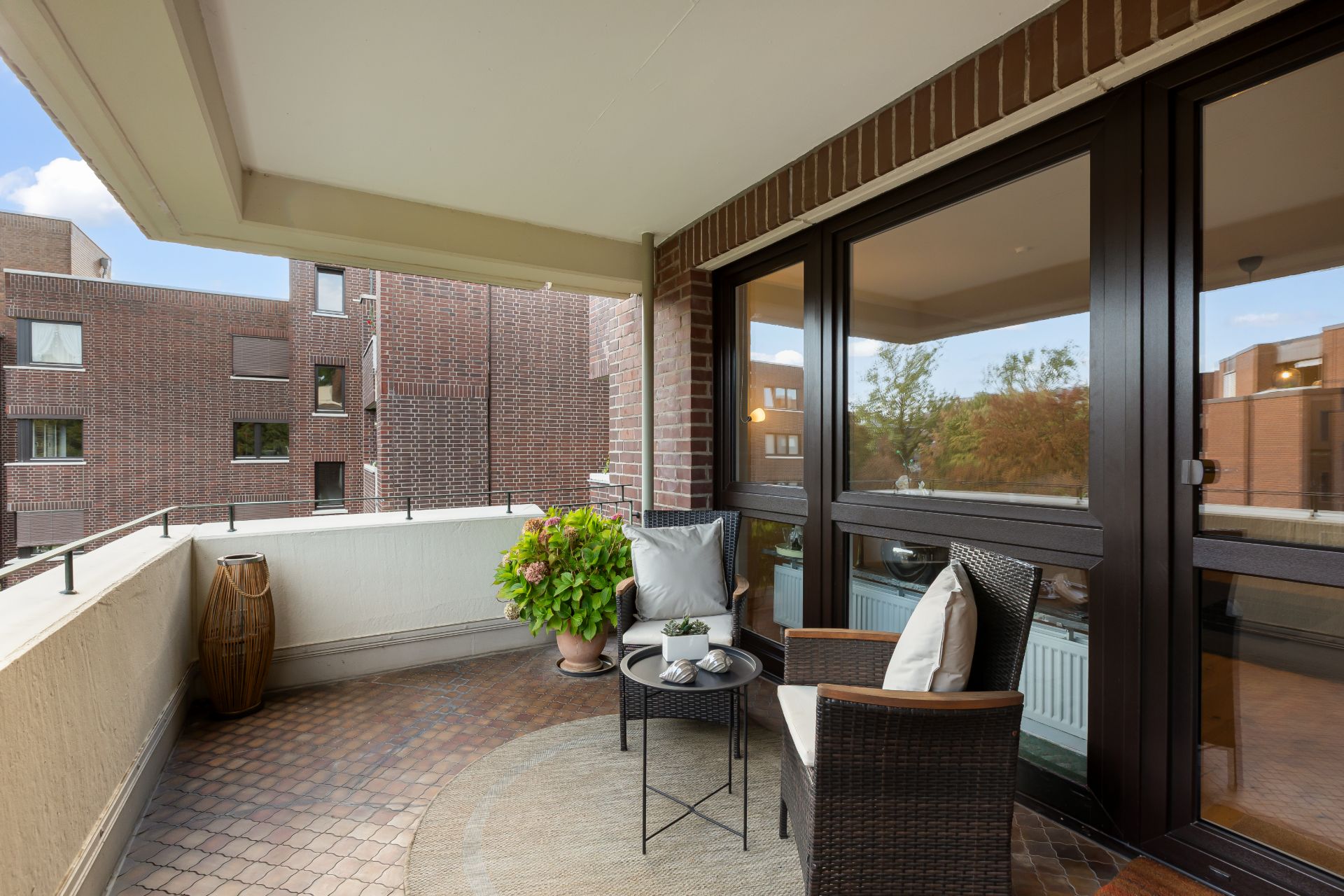 Überdachter Balkon mit zwei Stühlen, kleinem Tisch und Pflanze, angrenzend an große Fenster.