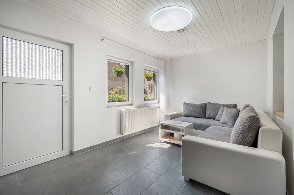 Helles Wohnzimmer mit grauem Ecksofa, Couchtisch und zwei Fenstern mit Blick nach draußen.