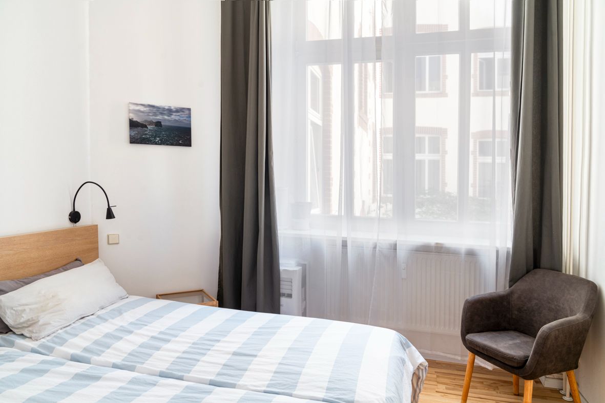 Helles Schlafzimmer mit Doppelbett, grauem Sessel und großem Fenster mit Vorhängen.
