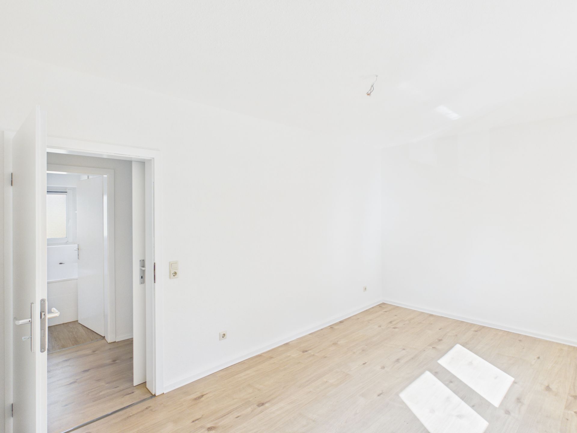 Heller leerer Raum mit Holzboden und weißen Wänden, offene Tür zu einem weiteren Zimmer.