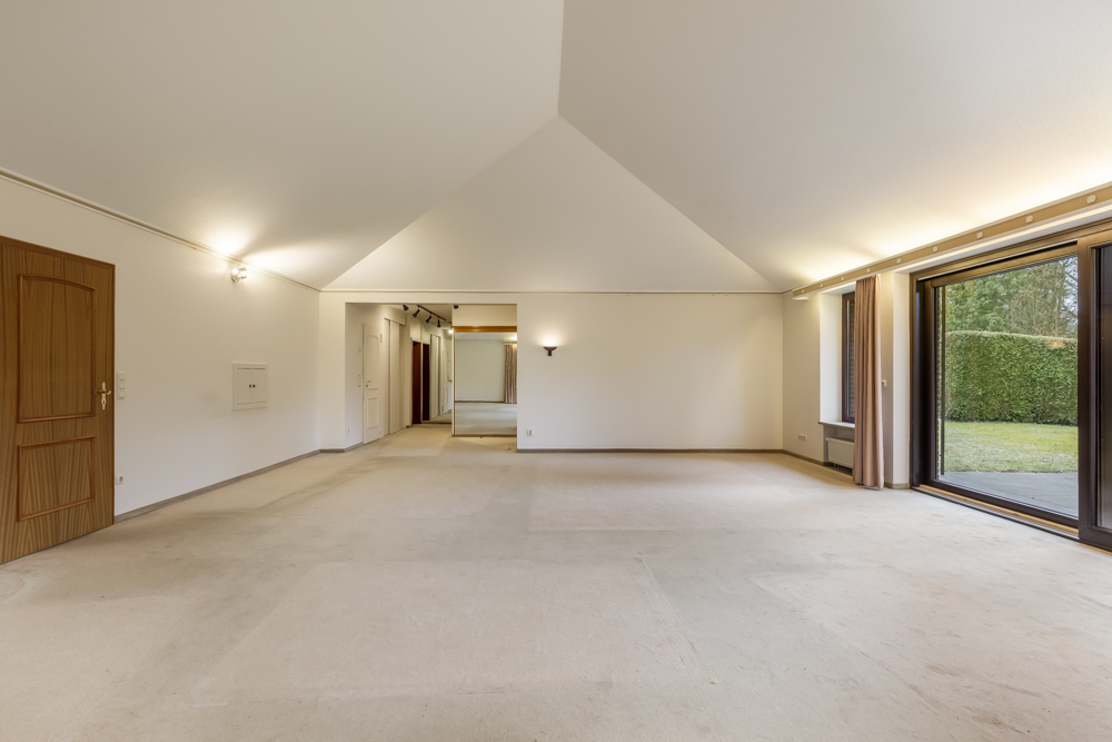 Geräumiges, leeres Wohnzimmer mit Teppichboden, großen Fenstern und Blick nach draußen auf einen Garten.