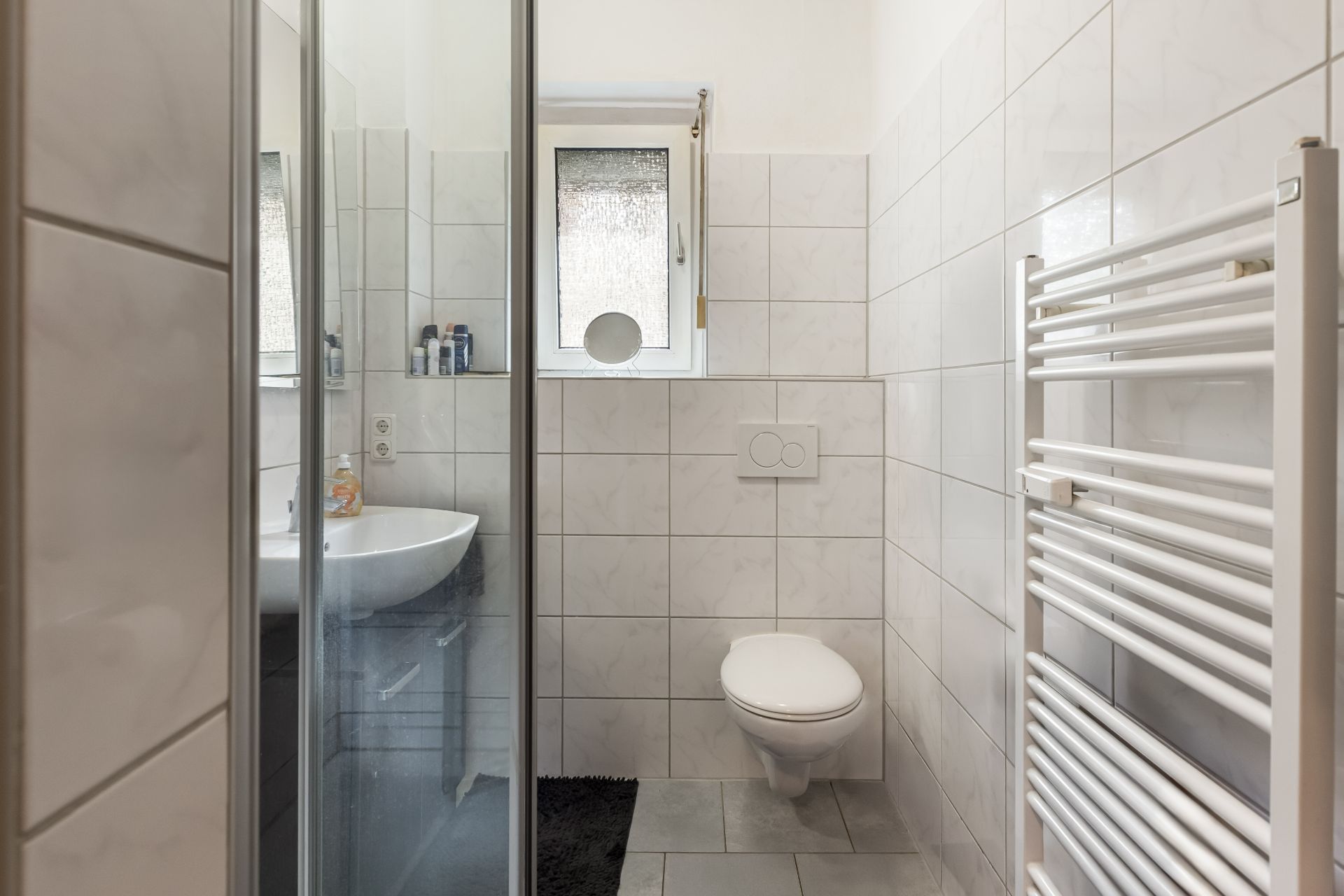 Kleines, gefliestes Badezimmer mit Dusche, Waschbecken, WC, Fenster und Handtuchheizkörper.