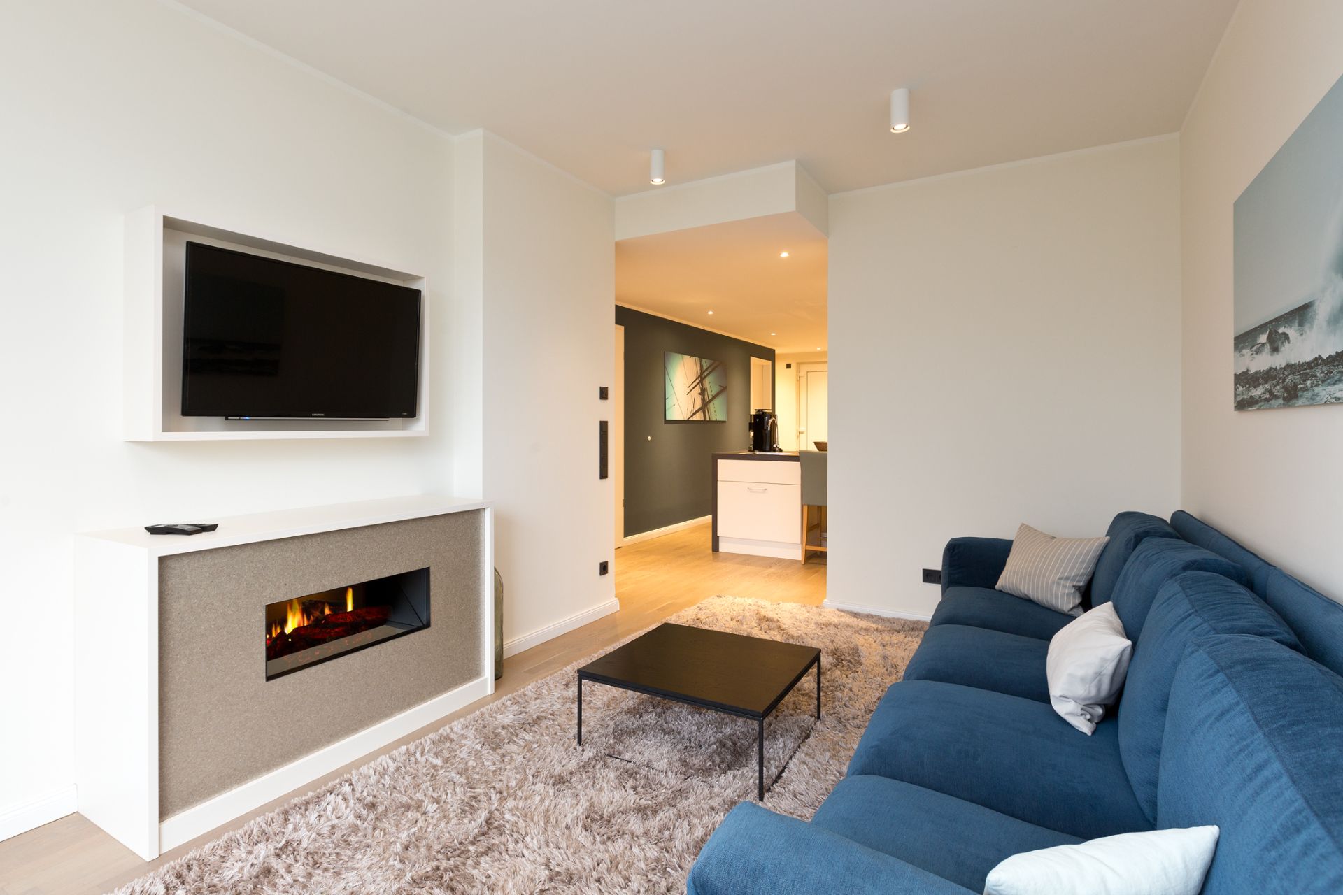 Modernes Wohnzimmer mit blauem Sofa, Couchtisch, Fernseher und Kamin, Blick zur offenen Küche.