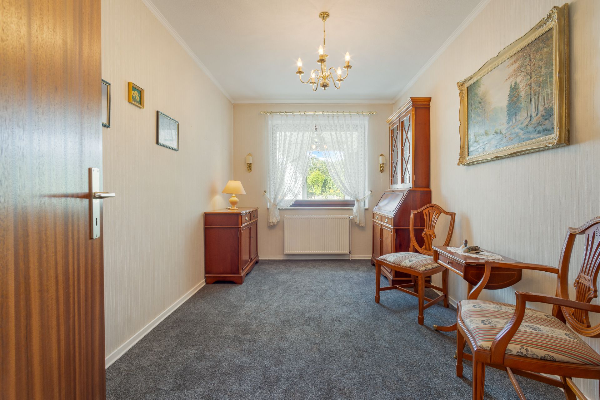 Helles Zimmer mit Teppich, Holzmöbeln, Fenster mit Gardinen und Kronleuchter.