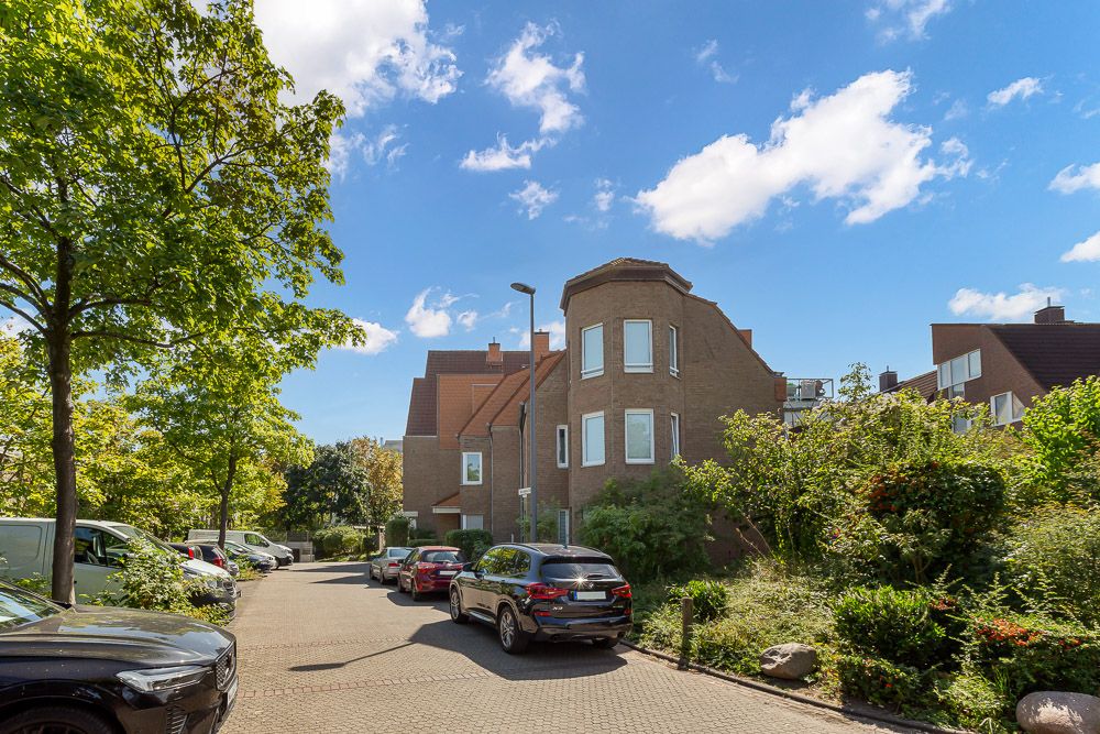 Wohnstraße mit geparkten Autos und einem großen Backsteinhaus unter blauem Himmel.