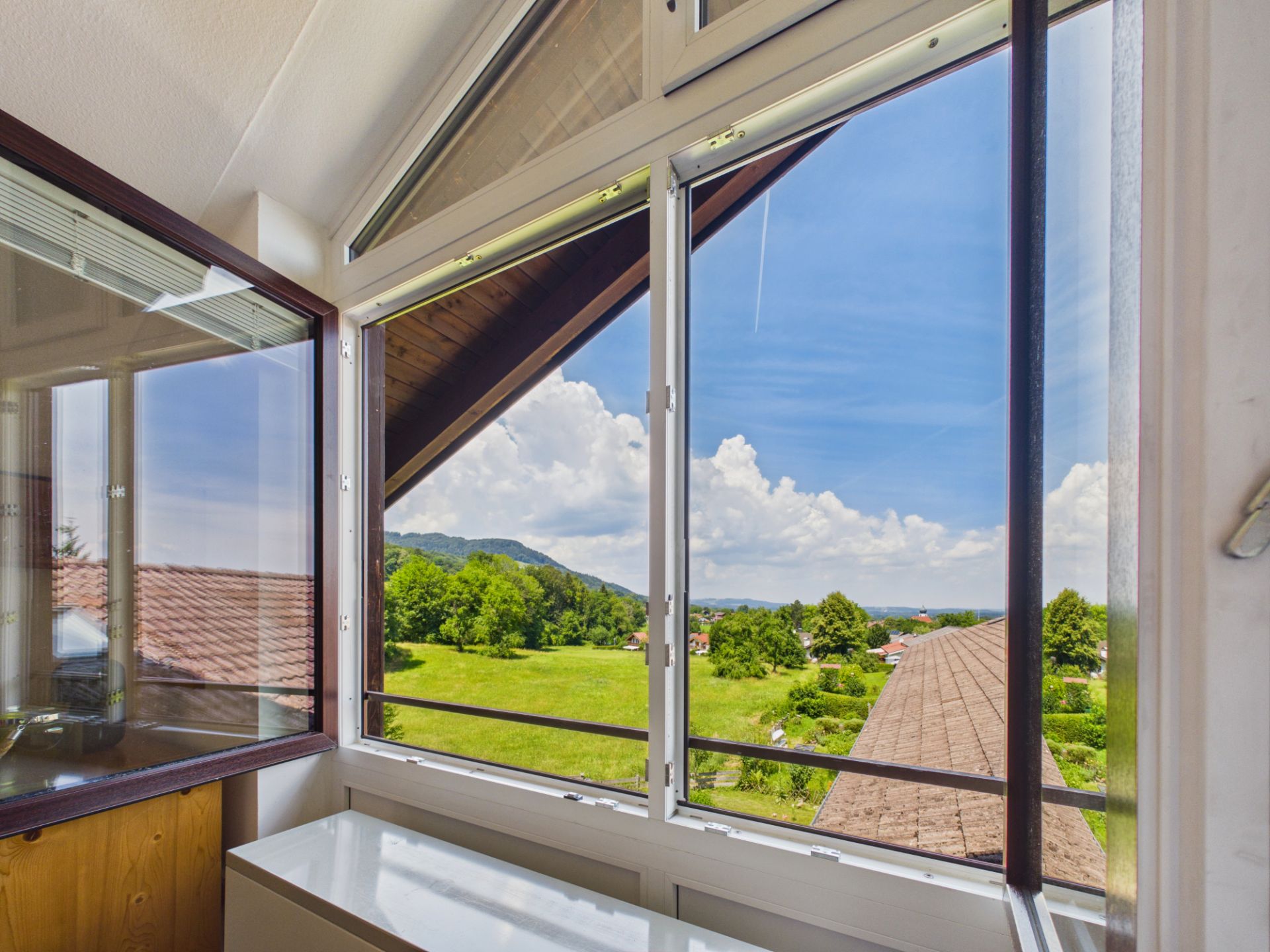 Blick aus einem offenen Fenster auf grüne Wiesen, Bäume und einen blauen Himmel mit Wolken.