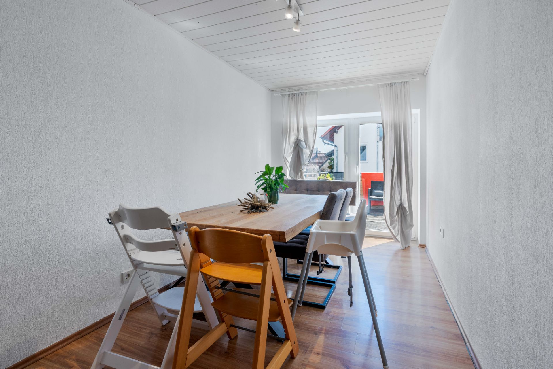 Esszimmer mit Holztisch, verschiedenen Stühlen und Pflanze, Tageslicht durch eine Glastür.