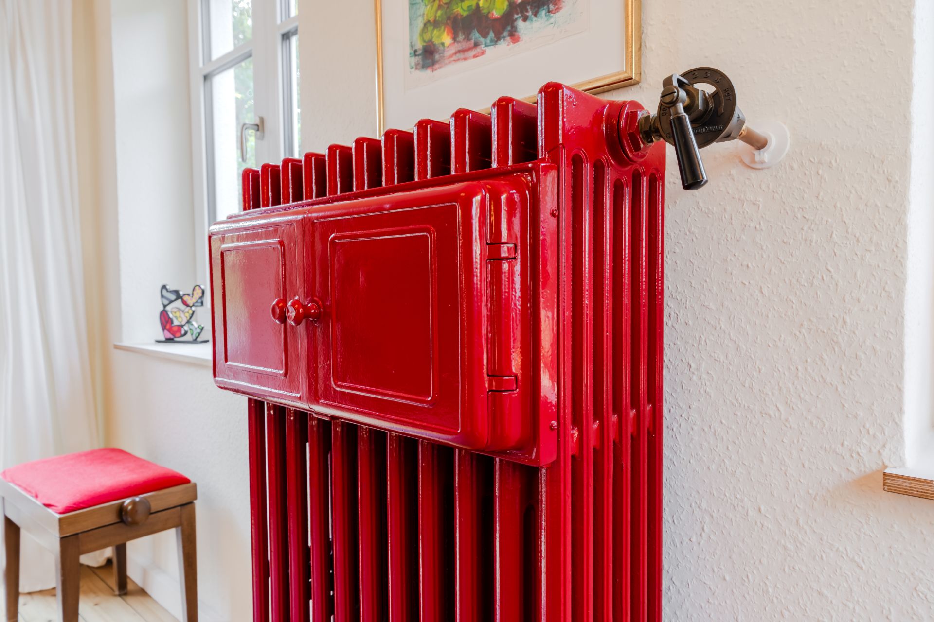 Roter Heizkörper mit Ventil im Raum neben einem Fenster und einem Stuhl mit rotem Sitzpolster.