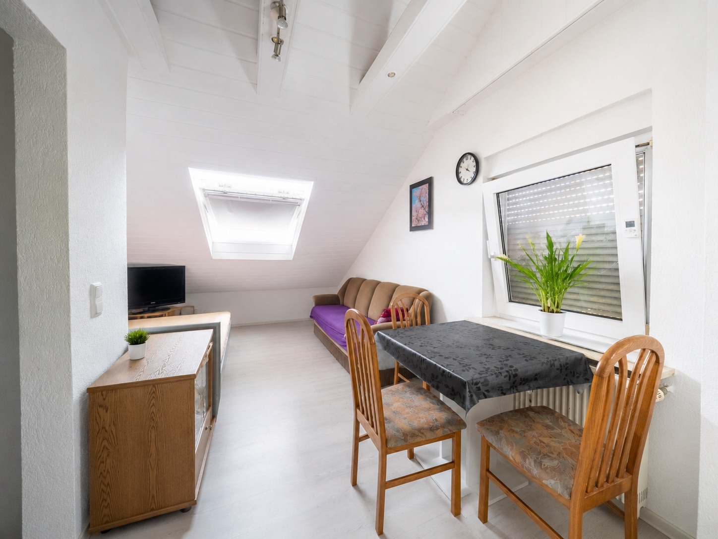 Helles Wohnzimmer mit Sofa, Esstisch, zwei Stühlen, Fernseher, Fenster und Pflanze.
