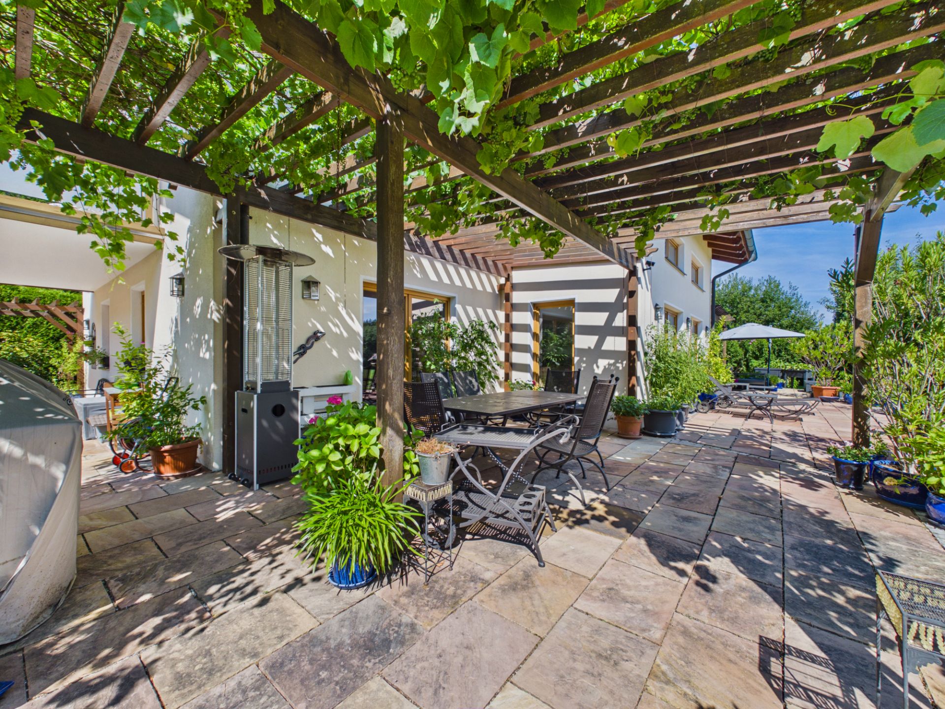 Große Terrasse mit Pergola, vielen Pflanzen und einem Esstisch mit Stühlen im Garten.