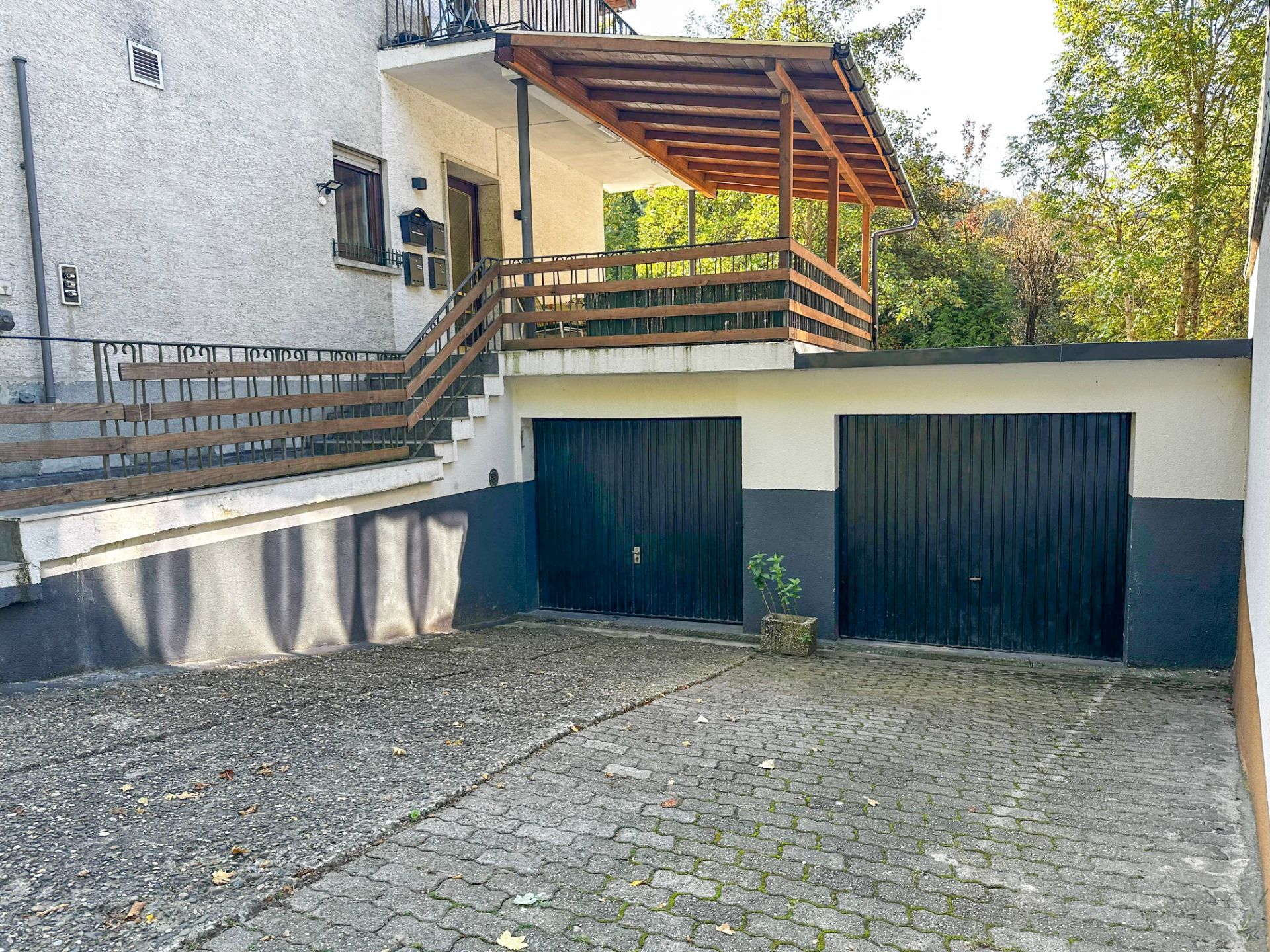 Zwei Garagentore unter einer überdachten Terrasse mit Geländer und Treppe, davor gepflasterter Hof.