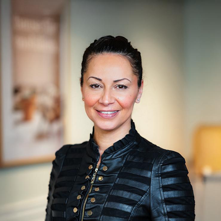 Frau mit dunkler Jacke steht in einem hellen Innenraum und lächelt in die Kamera.