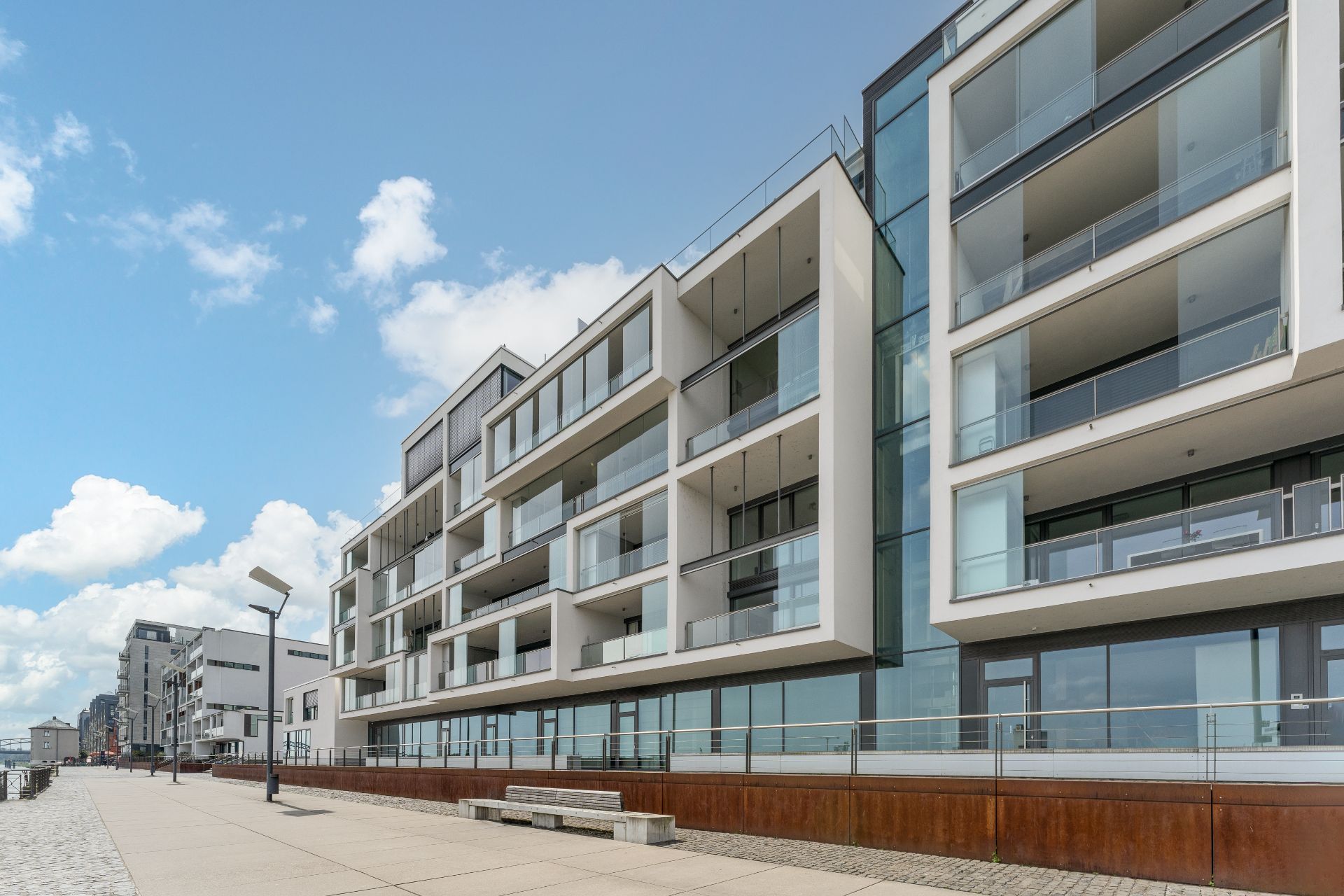 Moderne Wohngebäude mit großen Glasbalkonen an einer breiten Promenade bei blauem Himmel.