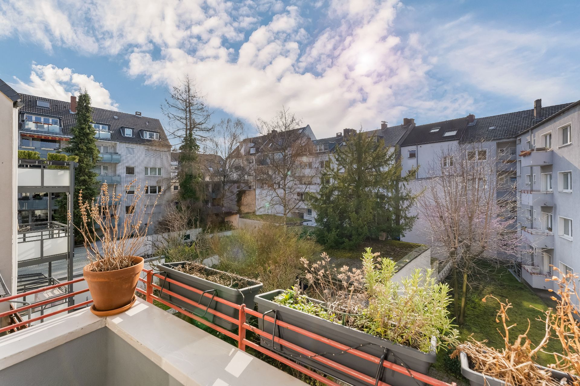 Blick von einem Balkon auf einen begrünten Innenhof mit mehreren Wohnhäusern und Pflanzenkästen am Geländer.