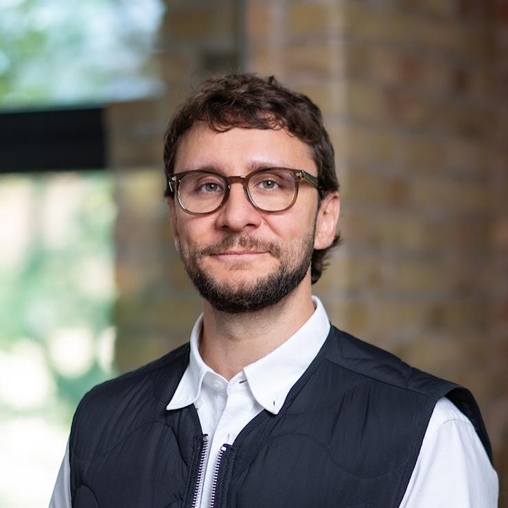 Ein Mann mit Brille und blauem Hemd steht in einem Raum mit Ziegelwand.