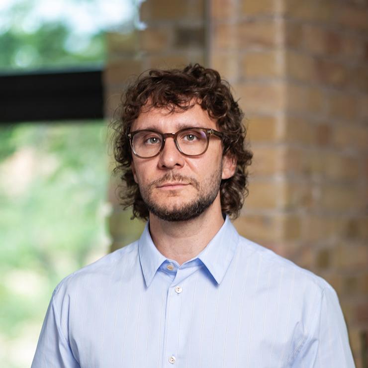 Ein Mann mit Brille und blauem Hemd steht in einem Raum mit Ziegelwand.