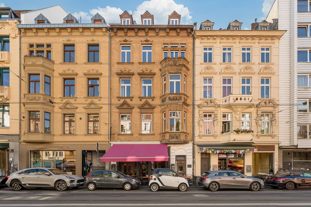Drei mehrstöckige Wohnhäuser mit großen Fenstern und Parkplätzen davor an einer Straße.