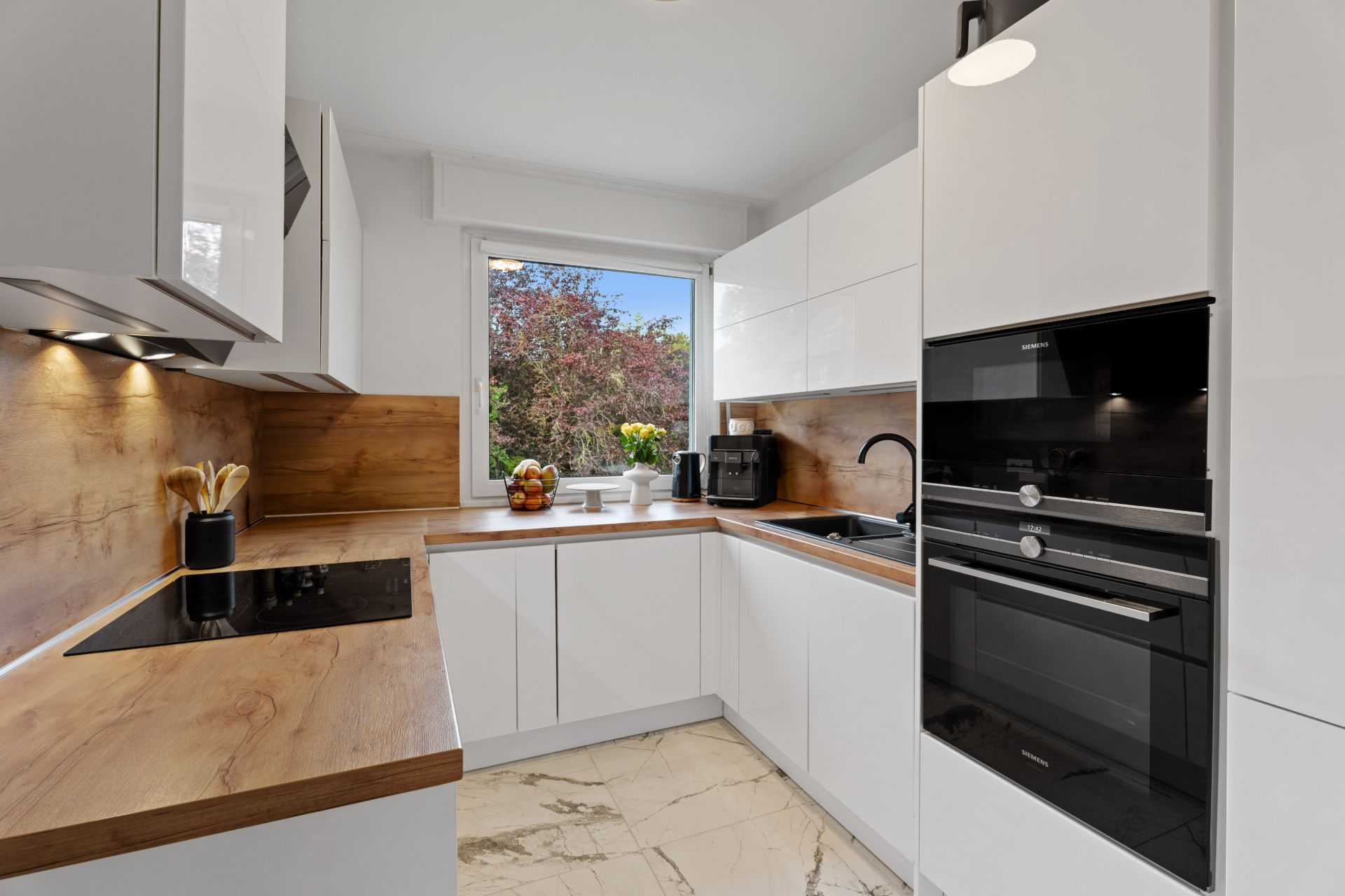 Moderne Küche mit weißen Schränken, Holzarbeitsplatten und großem Fenster mit Blick ins Grüne.