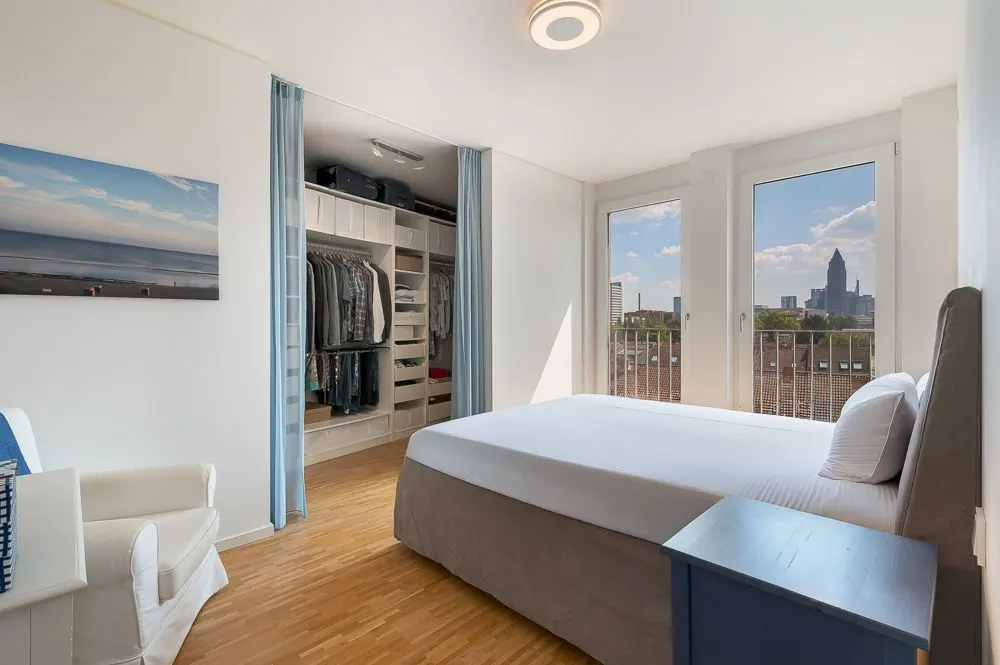 Helles Schlafzimmer mit Doppelbett, Sessel und offenem Kleiderschrank, große Fenster mit Stadtblick.