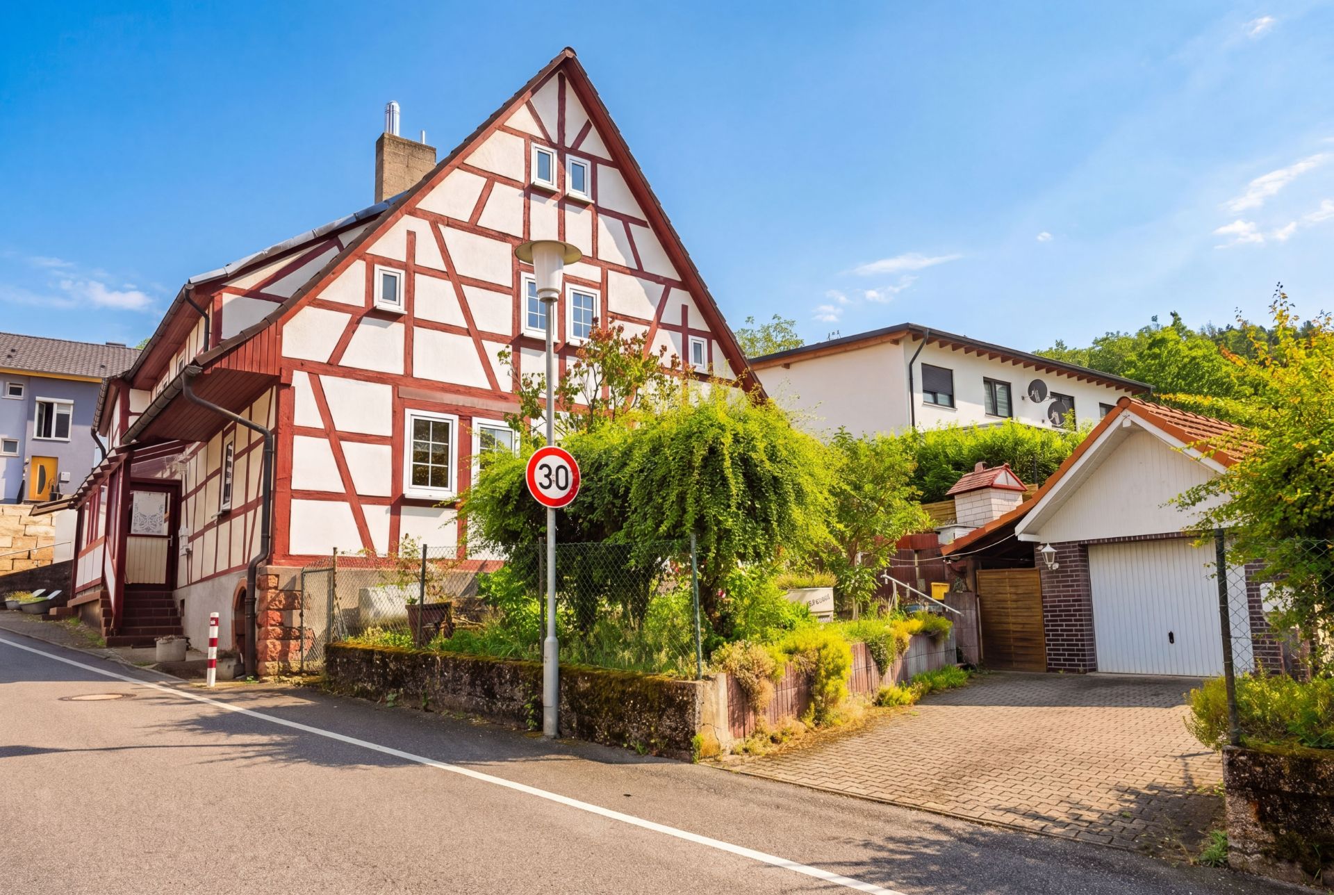 Fachwerkhaus mit Garten, Garage und 30 km/h Verkehrsschild an einer Straße bei sonnigem Wetter.