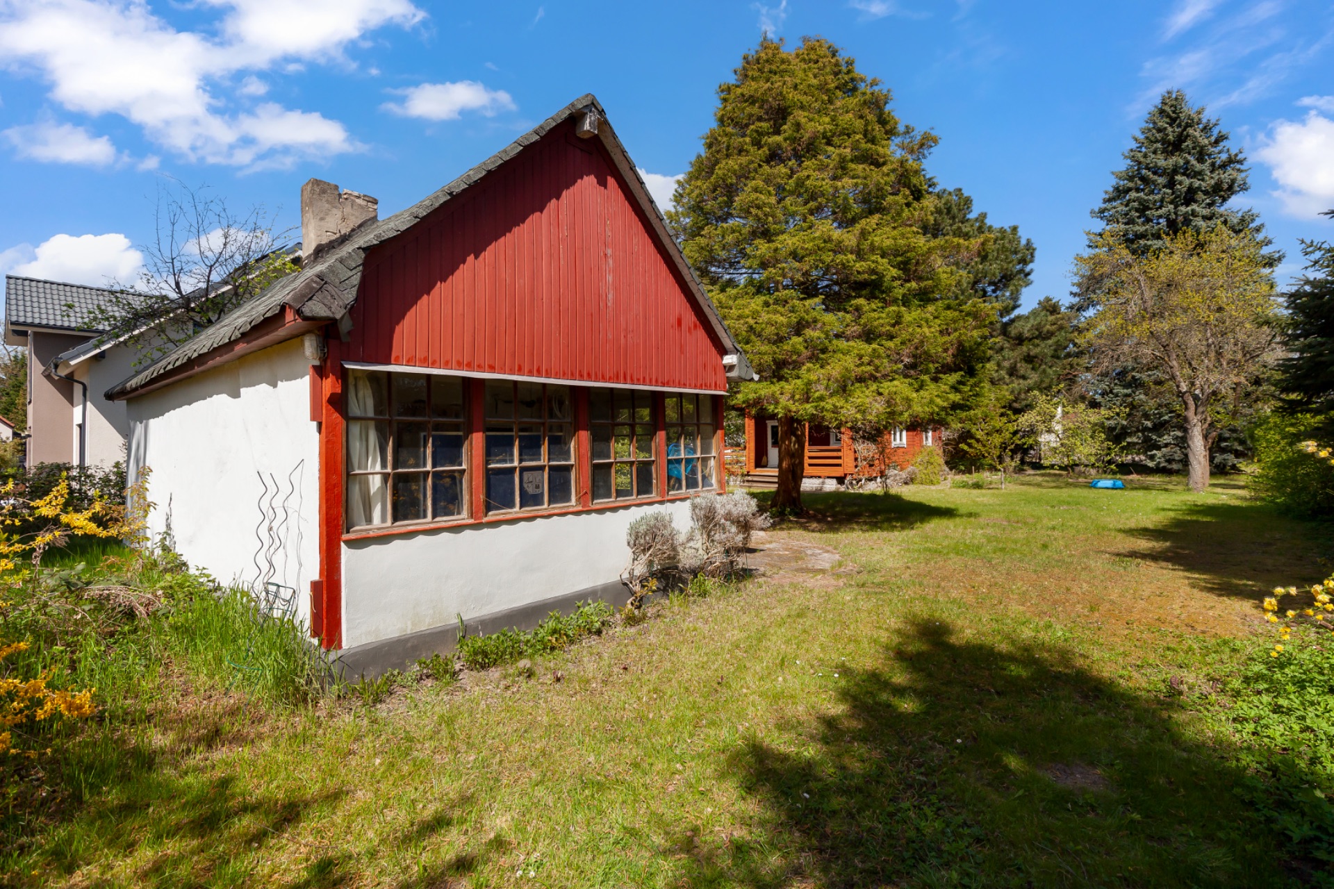 Kleines Haus mit roten Holzwänden und großem Garten, umgeben von Bäumen unter blauem Himmel.