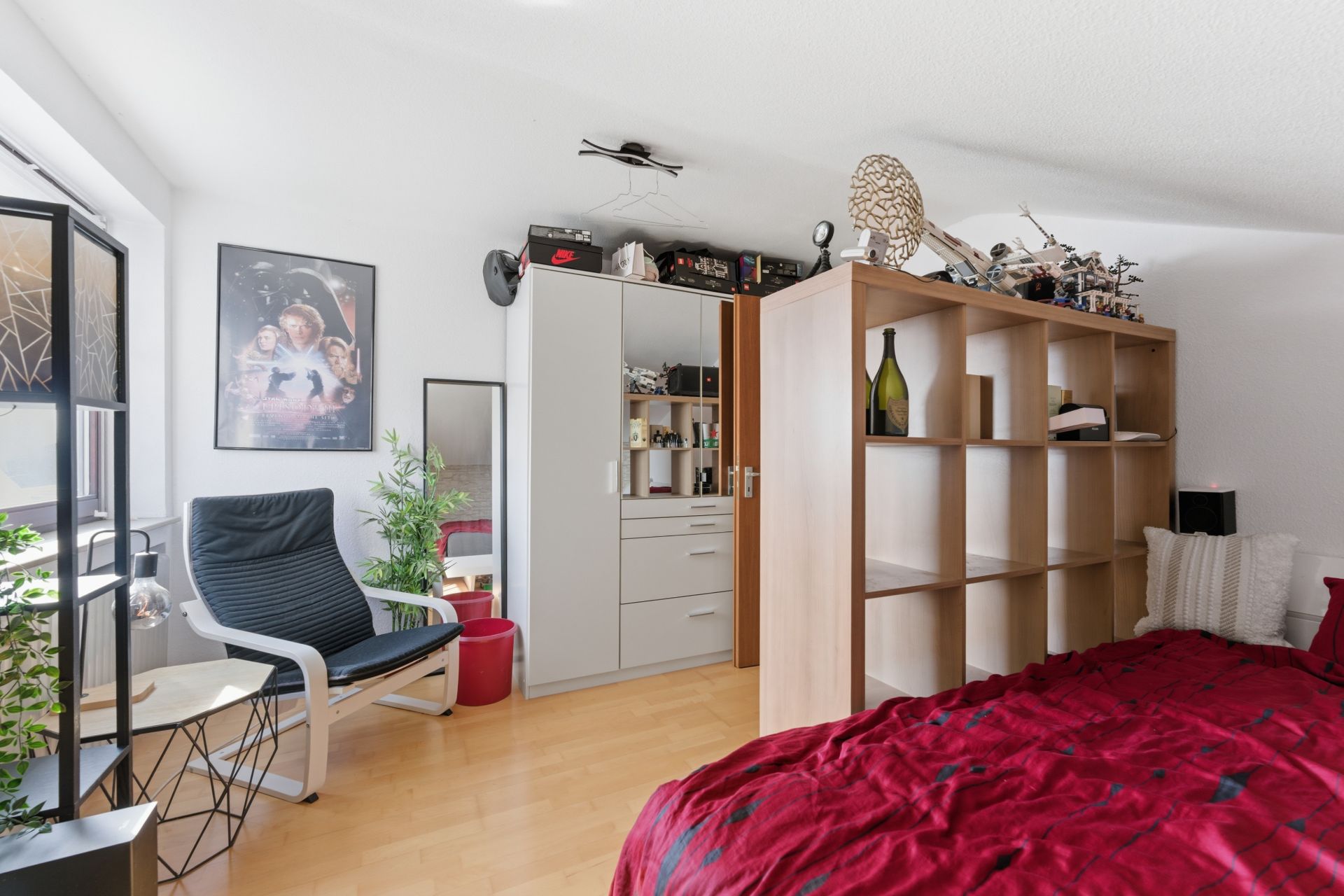 Helles Schlafzimmer mit rotem Bett, Regal als Raumteiler, Sessel, Schrank und Pflanzen.