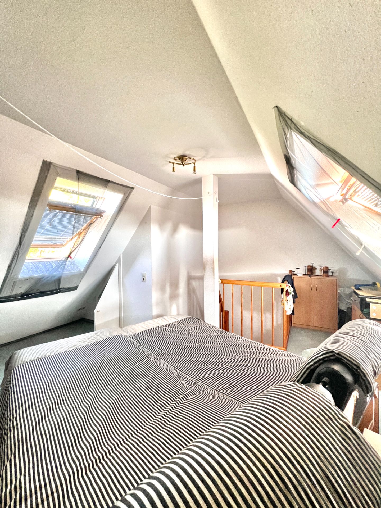Dachzimmer mit Bett, zwei Fenstern, Kommode und Treppengeländer.