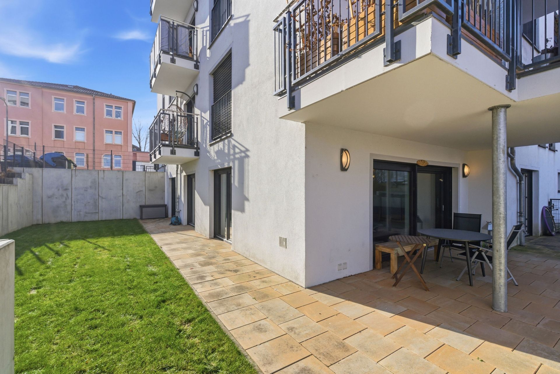 Terrasse mit Tisch und Stühlen, Rasenfläche und modernem Mehrfamilienhaus im Hintergrund.