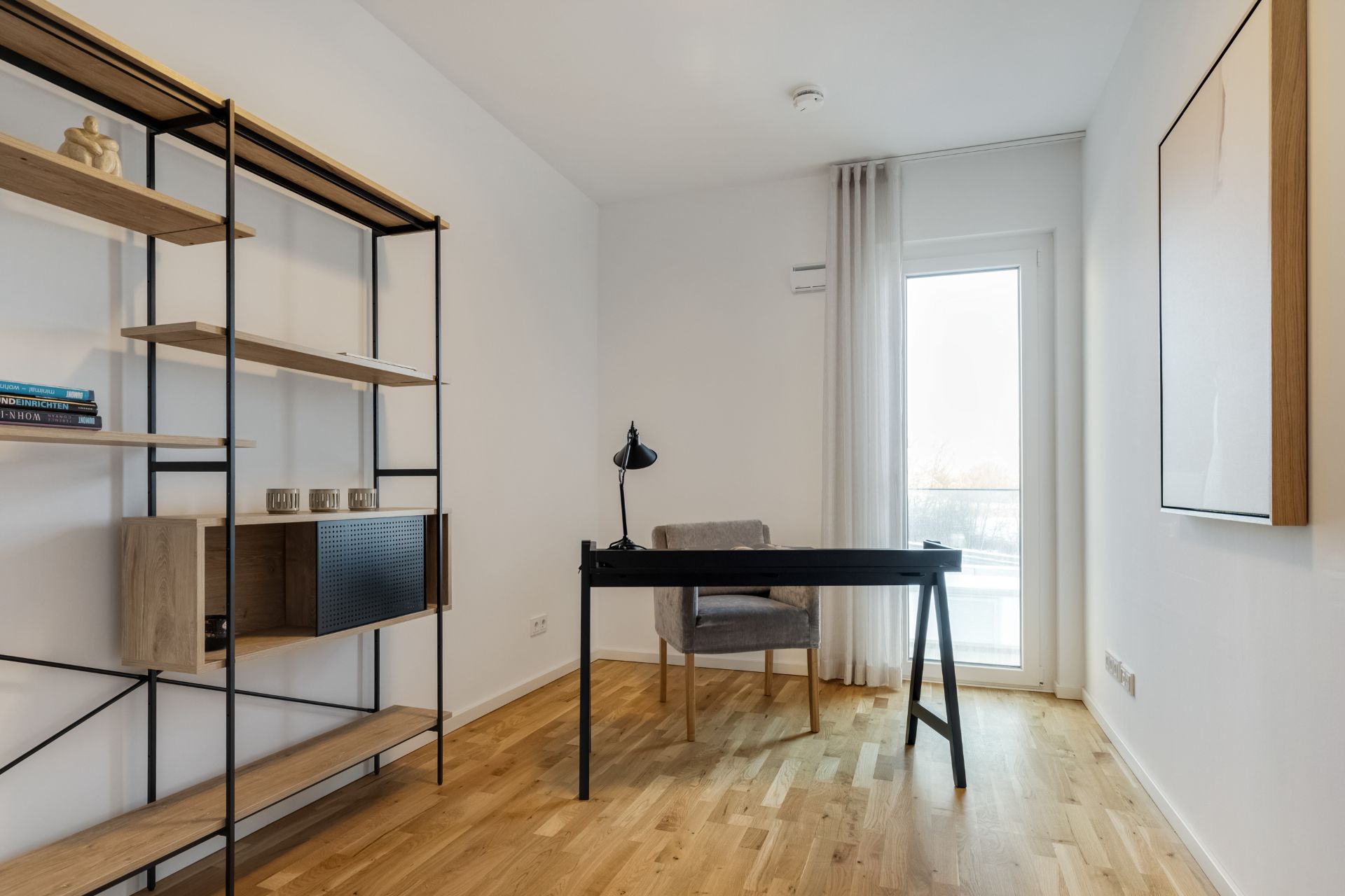 Helles Arbeitszimmer mit Schreibtisch, Stuhl, Regal und Fenster mit Vorhang.
