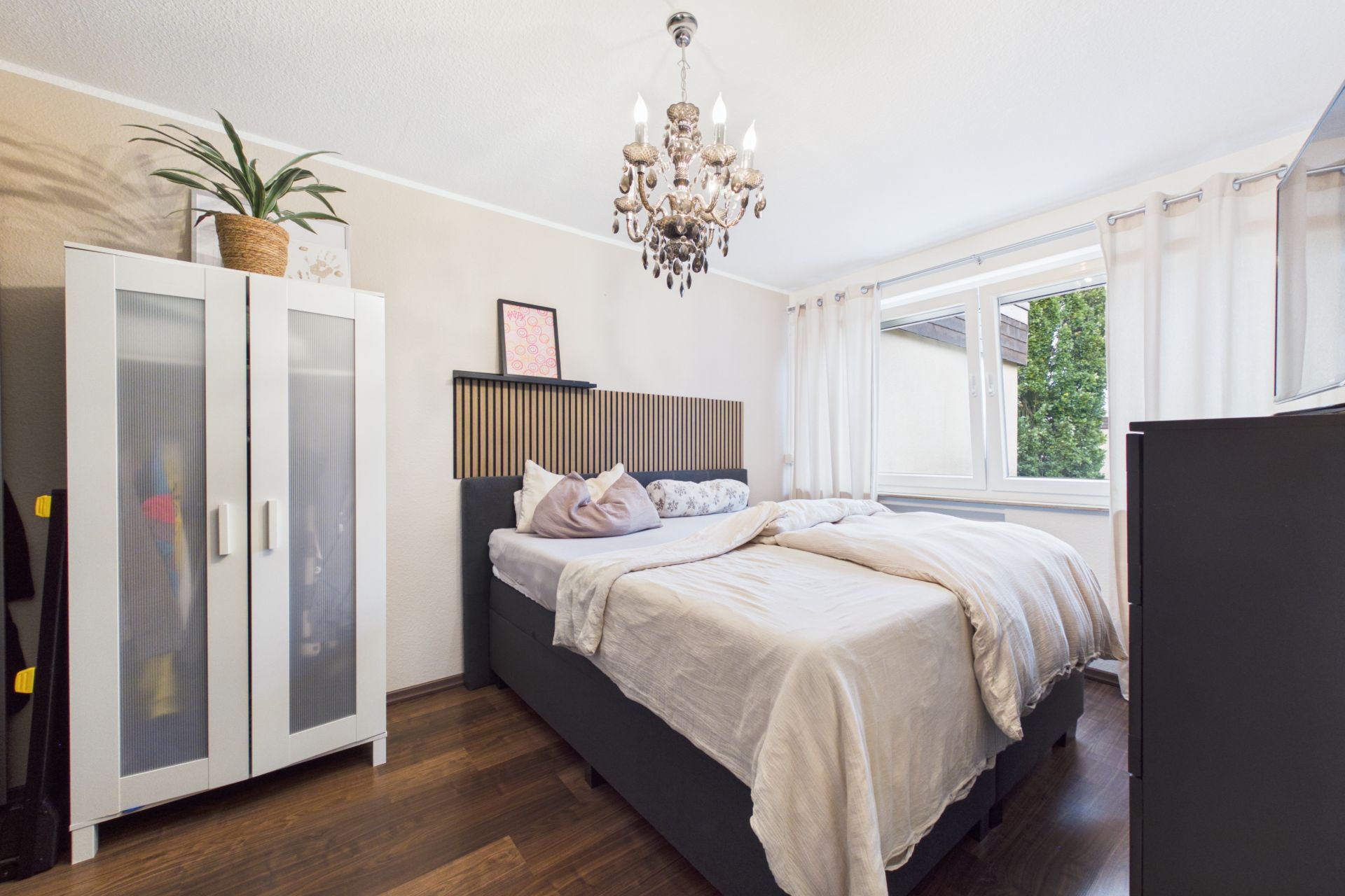 Schlafzimmer mit Doppelbett, weißem Kleiderschrank, Kronleuchter und großem Fenster mit hellen Vorhängen.